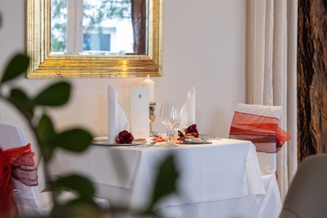 Ein romantisch gedeckter Esstisch für zwei Personen mit weißer Bettwäsche, roten Akzenten, einer brennenden Kerze und Rosenblättern erwartet Sie in einem eleganten Hotelzimmer, das mit einem goldgerahmten Spiegel und sanfter Beleuchtung geschmückt ist.